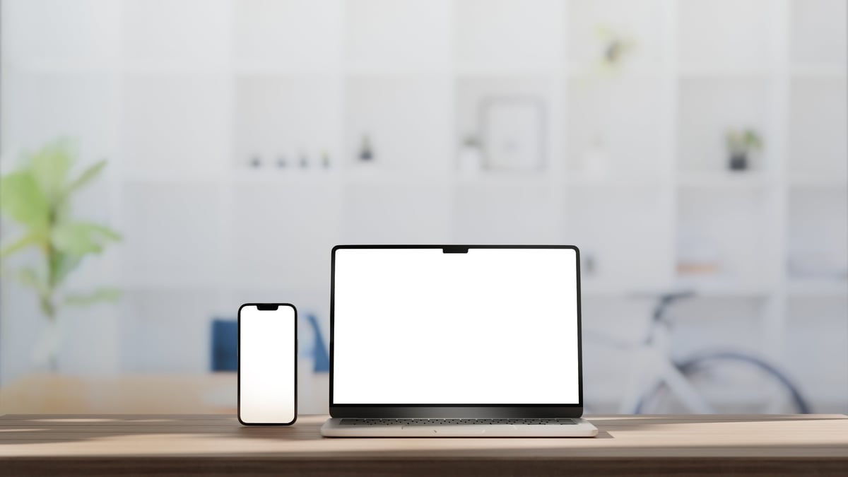 Laptop and smartphone with blank white screens on a wooden desk in a bright office.