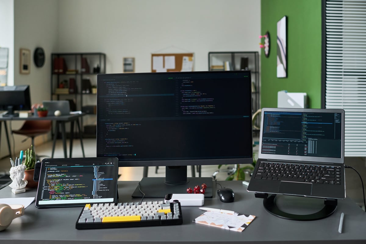 Wide shot of software development workspace displaying code on desktop monitor, laptop, tablet. Supporting programming, app creation, cybersecurity, tech marketing content